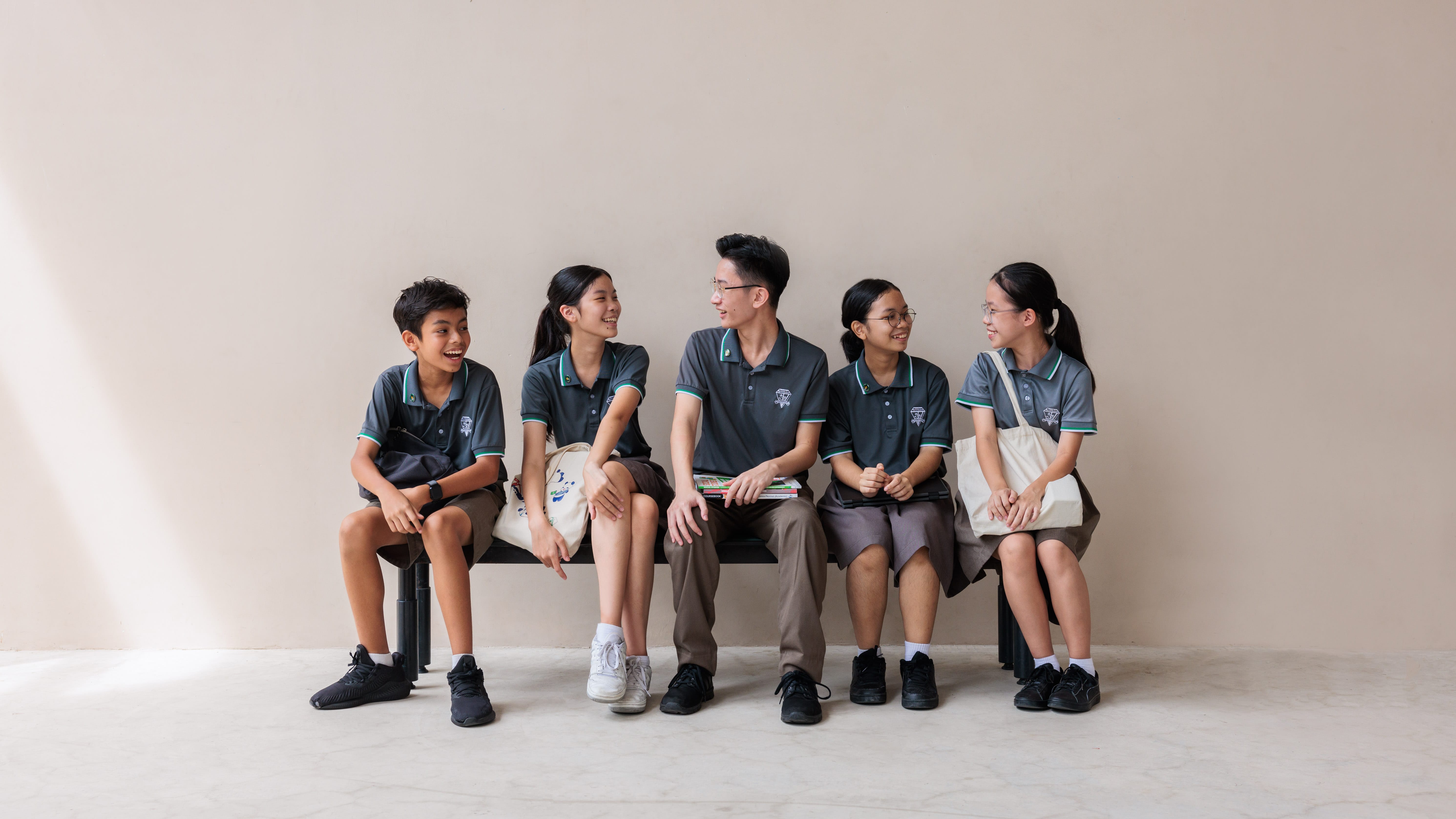 Five students in matching dark grey polo uniforms sit on a bench, smiling and chatting against a beige wall.